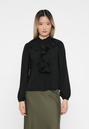 Blusa negra de manga larga con cuello acampanado y detalles en la parte delantera, que cuenta con cierre de botones y un tejido suave. Combinada con una falda verde.