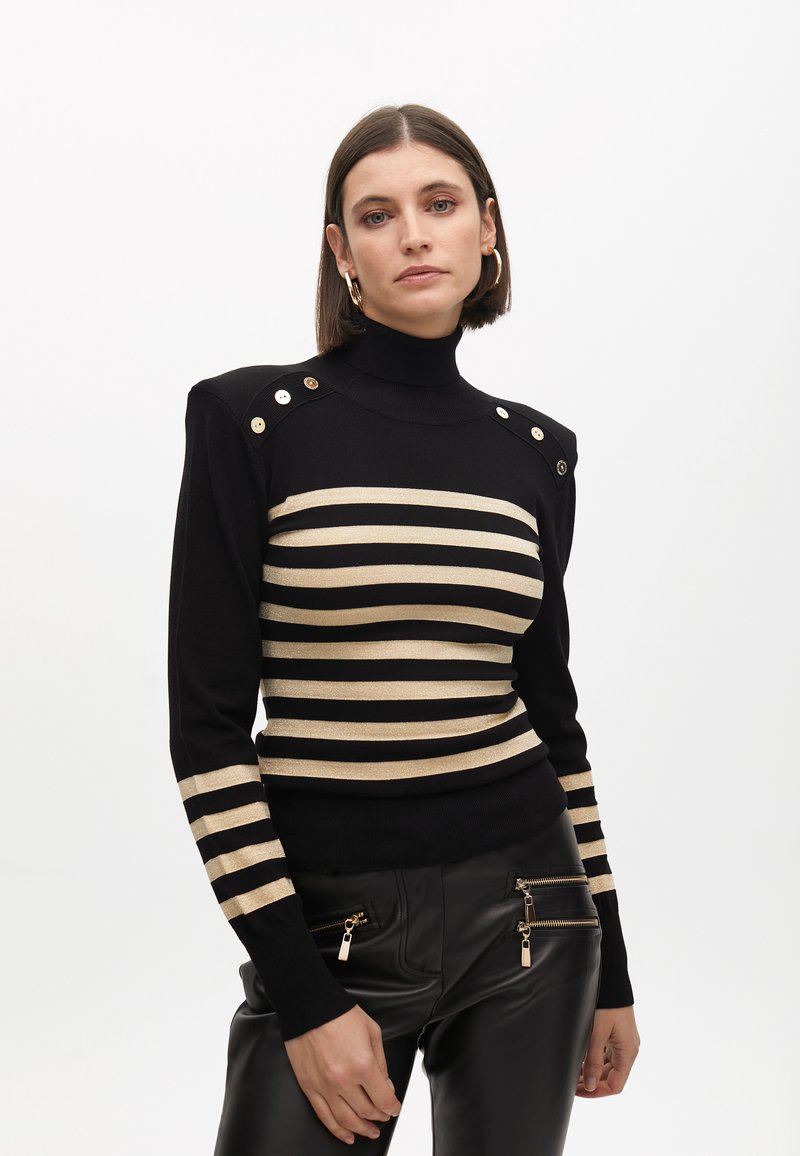 Crni i zlatni prugasti pleteni pulover s visokim vratom, dugmadima na ramenima i uskim krojem, u kombinaciji s crnim hlačama od umjetne kože s patentnim zatvaračima.