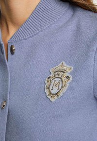 Cardigan lavorato a maglia di un leggero viola con bottoni argentati e una distintiva toppa ricamata in oro e nero a forma di stemma sul petto.