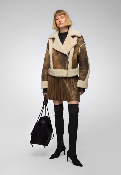 Bruine en crèmekleurige schapenvachtjas met een cropped ontwerp, gecombineerd met een geplooide bruine leren rok en zwarte laarzen tot boven de dij. Zwarte tas als accent.