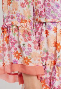 Květinové šaty s bledě růžovou látkou, na které jsou oranžové a fialové květy, volánovými rukávy a broskvovými akcenty na manžetách a lemu.