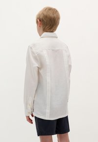 Witte linnen overhemd met lange mouwen en een klassieke kraag, voorzien van knoopmanchetten en verticale naden op de achterkant. Draagt bij donkere shorts.