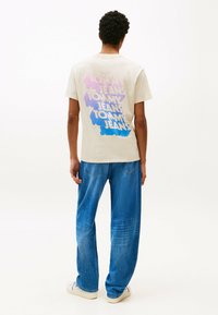 Beige bomulds t-shirt med multifarvet grafisk tekst "TOMMY JEANS" på ryggen, parret med løse blå denim jeans og hvide sneakers.