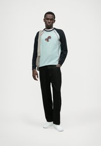 Γαλάζια φούτερ μπλούζα με μαύρες ράγες, κεντητό λογότυπο, κόκκινες λεπτομέρειες, συνδυασμένη με μαύρο παντελόνι και λευκά sneakers, κρατώντας τσάντα tote.