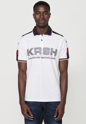 Bjela polo majica s tamnoplavim obrubom ovratnika i rukava, s izraženim logom "KRSH" i crvenim detaljima na rukavima. Nosila se s tamnim trapericama.