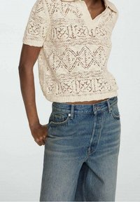 Haut tricot crème à manches courtes avec un col en V, présentant un design ajouré. Assorti à un jean en denim bleu taille haute.