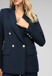 Veste bleu marine croisée avec revers crantés, boutons dorés et deux poches avant. Présente une coupe structurée et une texture de tissu lisse.