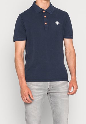 Man wearing a navy blue polo shirt with orange buttons and a small white logo on the chest, paired with light gray jeans.