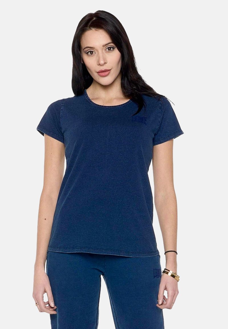 Jeune femme aux longs cheveux foncés portant un t-shirt bleu marine uni à manches courtes et un pantalon assorti sur fond blanc.