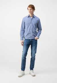 Blå og hvit rutete skjorte med knapper, slim-fit denim jeans, hvite joggesko, modell som står mot en nøytral bakgrunn.