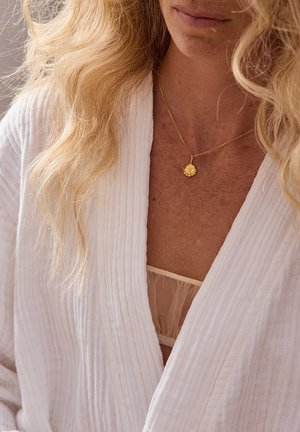Robe blanche texturée avec un design à côtes subtil, assortie d'un délicat collier en pendentif doré avec un charme circulaire. Les cheveux semblent ondulés.