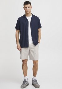 Marineblauwe kortemouwen overhemd met knopen, gedragen over een wit T-shirt. Beige shorts, grijze sneakers met accenten. Eenvoudig ontwerp en casual pasvorm.