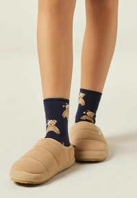 Tan corduroy slip-on slippers med en tekstureret overflade og gummisåler, parret med marineblå sokker med bjørnedesigns iført julestræhatte.