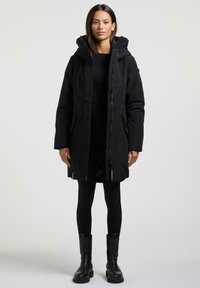 Zwarte lange jas met capuchon en ritsluiting, met twee voorzakken en een toggledrawstring. Draagt over zwarte leggings en laarzen.