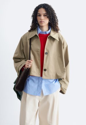 Personne aux cheveux bouclés porte une veste courte beige, un pull rouge, une chemise bleue à boutons, un pantalon crème et porte un sac à épaule foncé devant un fond uni.
