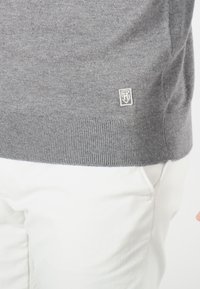 Pull en maille gris avec un ourlet et des poignets côtelés, présentant un petit logo brodé en blanc en bas à gauche. Associé à un pantalon blanc.