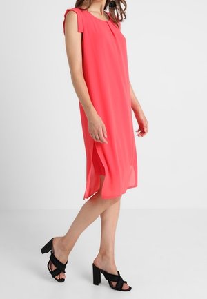 Mujer con un vestido midi coral sin mangas con aberturas laterales y sandalias negras de tacón alto, posando frente a un fondo liso.