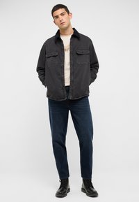 Czarna kurtka z tkaniny z zapięciem na guziki, dwoma kieszeniami na piersiach i kołnierzem; noszona na kremowej koszulce i zestawiona z ciemnymi dżinsami oraz czarnymi butami.