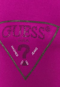 Top fucsia Guess con decorazioni in strass, caratterizzato da un logo triangolare e dal testo "WASHED" e "JEANS" in caratteri in grassetto.