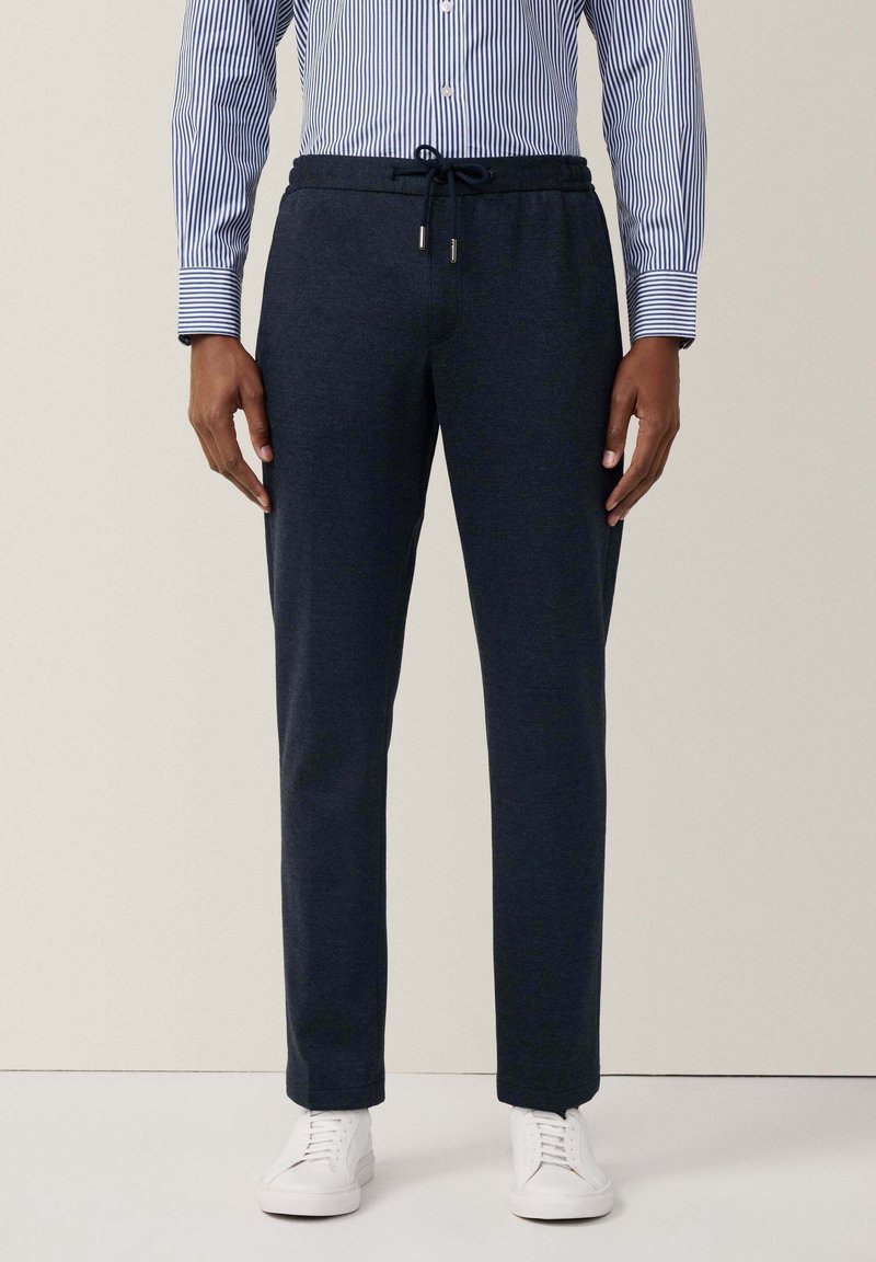Homme portant un pantalon bleu marine à cordon, des baskets blanches et une chemise rayée bleu et blanc à boutons, debout devant un fond uni.