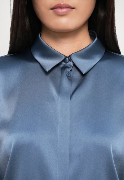 Primer plano de una persona que lleva una camisa de satén azul abotonada con un cuello puntiagudo y con el cabello oscuro largo visible.