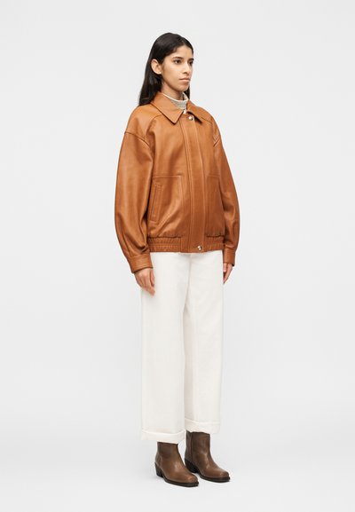 Γυναίκα που στέκεται φορώντας καφέ δερμάτινο μπουφάν, λευκό παντελόνι φαρδιάς γραμμής και καφέ μποτάκια μέχρι τον αστράγαλο, με φόντο απλό και φωτεινό.