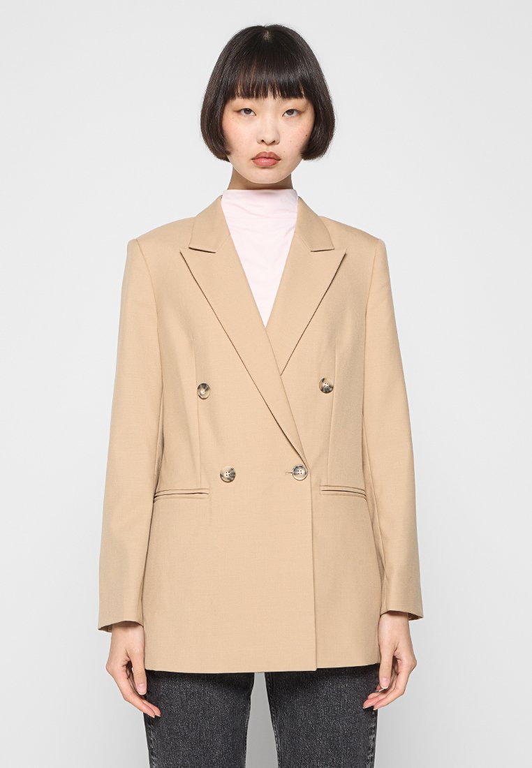 Beige doppelreihiger Blazer mit zwei Vorderaschen, silbernen Knöpfen und scharfen Revers, über einem hellrosa Rollkragenpullover.