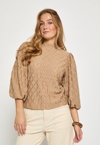 Beige tekstureret bluse med diamantmønstret strik, høj halsudskæring, pufærmer og kort skåret kant. Bær den med cremefarvede bukser og et armbånd.