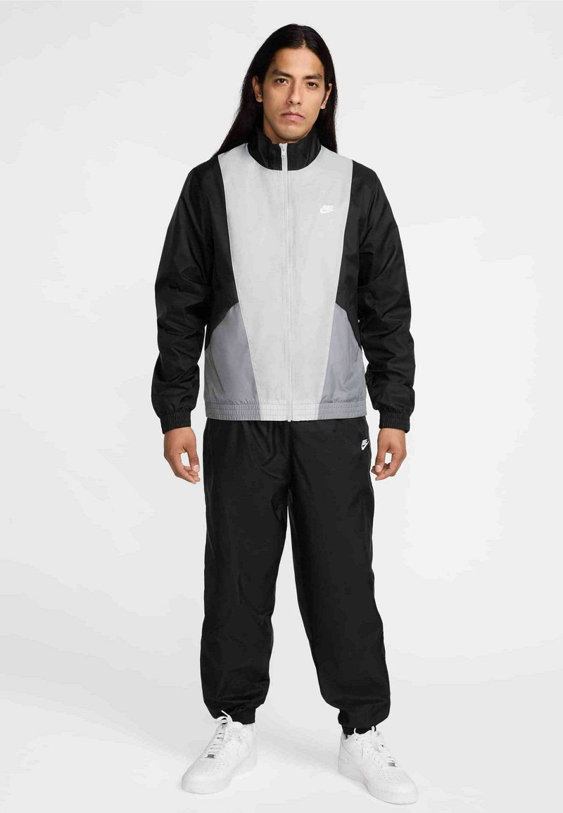 Αντιανεμικό μπουφάν σε μαύρο και γκρι, με φερμουάρ, ψηλό γιακά και ελαστικά μανίκια. Συνδυάζεται με αντίστοιχο μαύρο παντελόνι και λευκά αθλητικά παπούτσια.