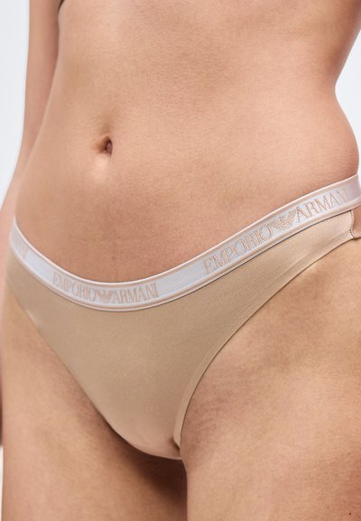 Κοντινό σε πρόσωπο που φοράει μπεζ εσώρουχα Emporio Armani με λευκή ζώνη μέσης με λογότυπο.