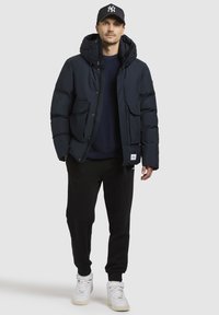 Marineblå pufferjakke med hætte, med lynlås og trykknapslukning, to frontlommer; parret med sorte sweatpants og hvide sneakers.