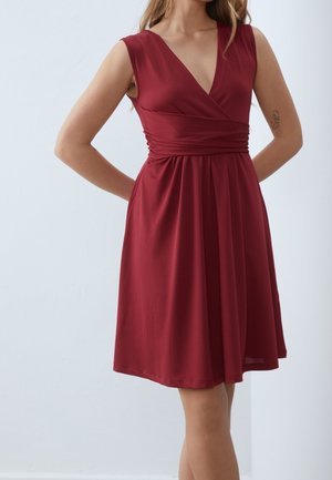Femme portant une robe sans manches, longueur genou, rouge profond avec col en V, taille cintrée et légers plis, debout les mains dans le dos.