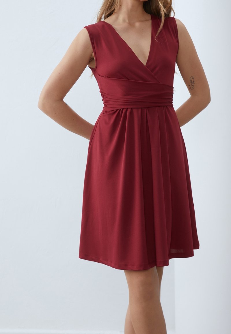 Femme portant une robe sans manches, longueur genou, rouge profond avec col en V, taille cintrée et légers plis, debout les mains dans le dos.