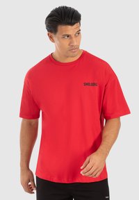 Rød bomulds t-shirt med afslappet pasform, korte ærmer, rund halsudskæring og et sort "SMILODON" logo på venstre bryst. Simpelt design.
