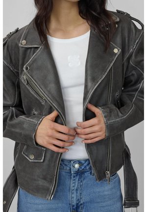 Personne portant une veste en cuir noire délavée par-dessus un t-shirt blanc côtelé et un jean bleu, boutonnant la veste à l'avant.