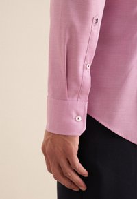 Camicia a maniche lunghe di colore rosa con texture, dotata di polsini abbottonati e con bottoni bianchi rifiniti in rosso, esposta su una mano contro uno sfondo neutro.