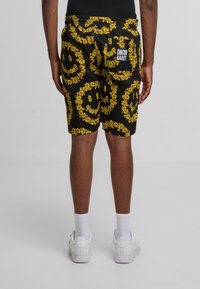 Sorte shorts med et gult blomster-smilansikt mønster, elastisk midjebånd, og en lomme med et logo. Vist fra baksiden.