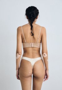 Bralette beige cropped et thong en satin, avec des bretelles ajustables et des coutures décoratives, vus de dos sur un fond clair.