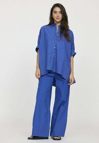 Chemise bleue à manches courtes, ample et boutonnée à l'avant, assortie à un pantalon bleu à jambes larges. Le tissu semble lisse et léger.