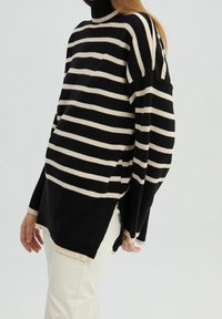 Femme portant un pull à col roulé oversize rayé noir et blanc avec des fentes latérales et un pantalon écru sur fond uni.