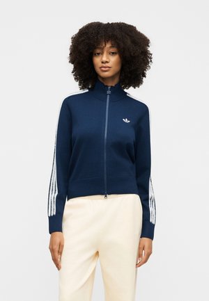 Mujer con cabello rizado lleva una chaqueta azul marino con cremallera y rayas blancas en las mangas y pantalones color crema, de pie contra un fondo liso.
