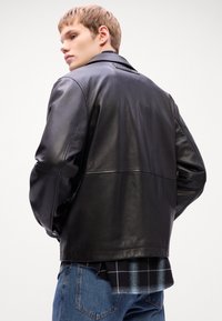 Black leather jacket with a smooth texture, featuring a classic collar and a back yoke. Worn over a checkered flannel shirt.