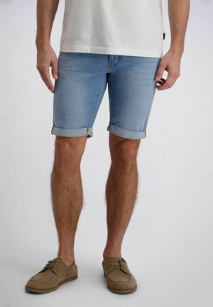Man die lichtblauwe opgerolde denim shorts, een wit t-shirt en beige casual schoenen draagt, staand tegen een eenvoudige lichte achtergrond.