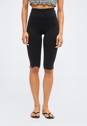 Γυναίκα με ψηλόμεσα μαύρα κολάν μέχρι το γόνατο με μικρές πλαϊνές σκισμές, μαύρα σανδάλια τύπου σαγιονάρα και γκρι κοντό τοπ.