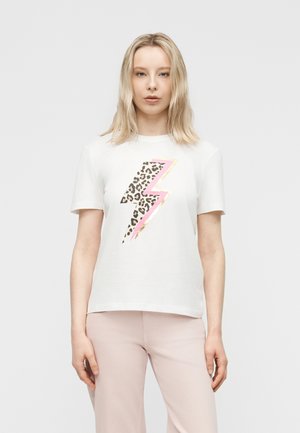 Mujer rubia con camiseta blanca con un rayo de estampado de leopardo delineado en rosa y oro, combinada con pantalones rosa claro, de pie vista frontal.