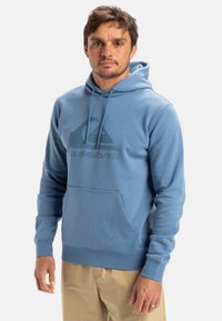Ljusblå hoodie med känguruficka, justerbar snörning och en mörk logotryck på framsidan. Mjuk tyg, avslappnad passform.