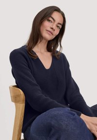 Strickpullover in Marineblau mit V-Ausschnitt und langen Ärmeln, versehen mit einem strukturierten Muster. Über einer dunklen Jeanshose getragen, sitzt auf einem Holzstuhl.