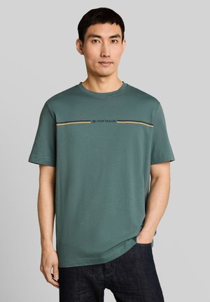 Mann trägt grünes kurzärmeliges Tom Tailor T-Shirt mit gelben und schwarzen Streifen über der Brust, kombiniert mit dunklen Jeans, steht vor grauem Hintergrund.
