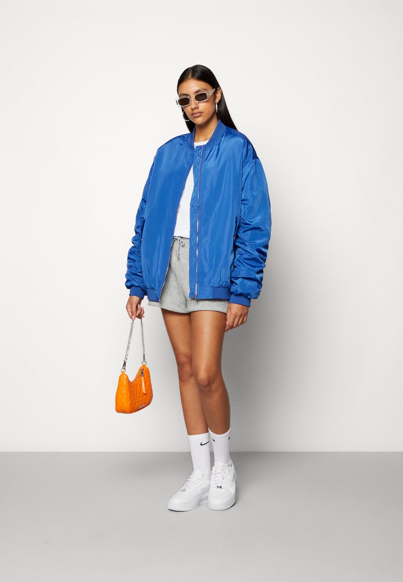 Veste bomber bleue, t-shirt blanc, shorts gris, sac à main orange, baskets blanches et lunettes de soleil. Tissu lisse, coupe décontractée et couleurs vives.