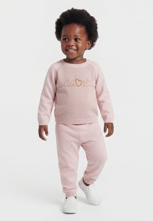 Smilende småbarn som har på seg en lys rosa genser med teksten "Adoré", matchende bukser og hvite slip-on sko, står foran en hvit bakgrunn.